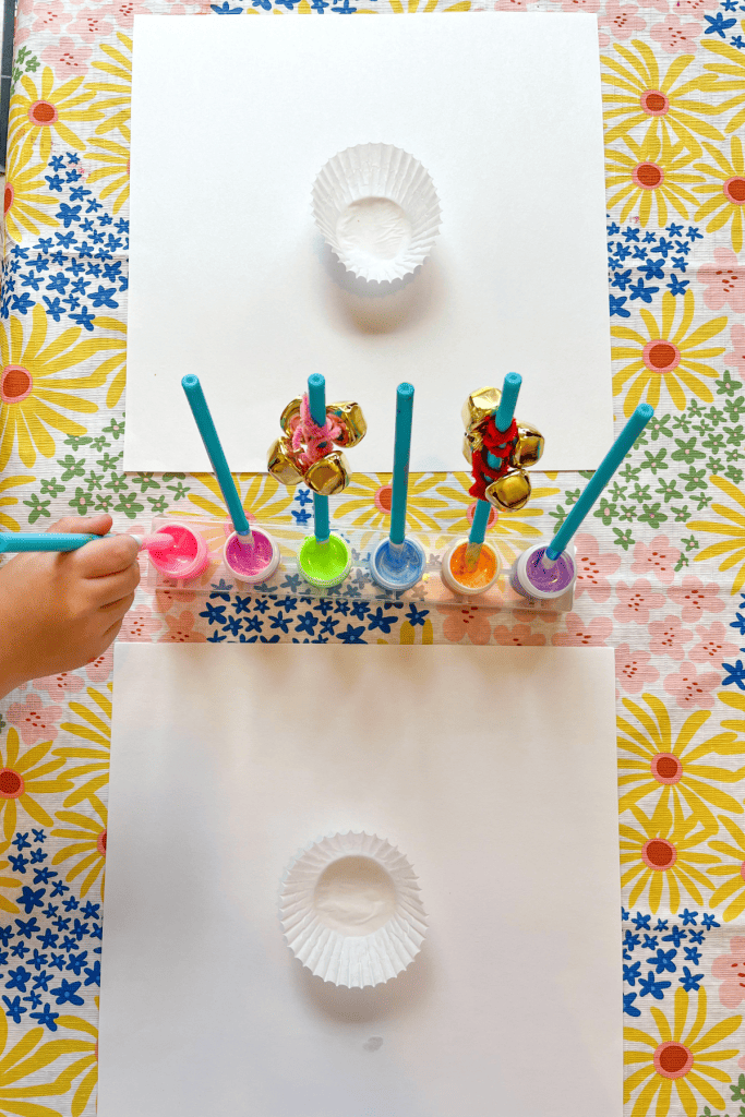 Two white papers with cupcake liners glued to the middle sit on both sides of a container with six paint wells filled with pastel paint and paintbrushes. 