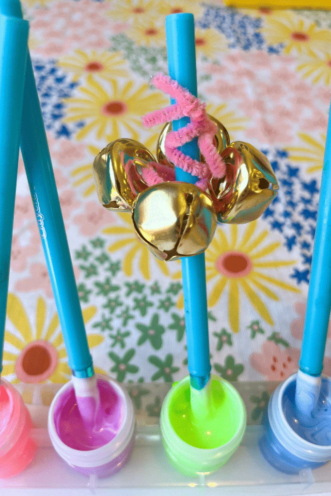 The top of a blue paintbrush has a pink pipe cleaner with four golden bells threaded onto it and is wrapped around the top of the paintbrush. 