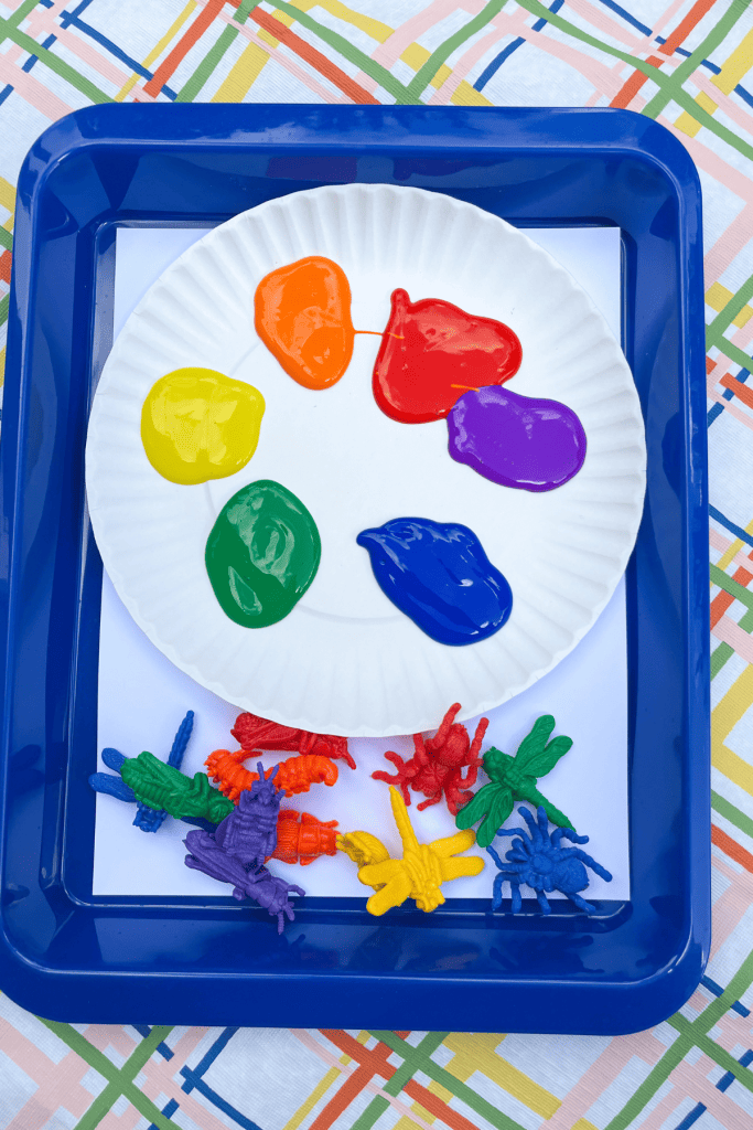 Materials for the project: Six puddles of paint (red, orange, yellow, green, blue, and purple) sit on a white paper plate, colorful plastic big toys, and white paper. All materials sit in a shallow blue tray. 