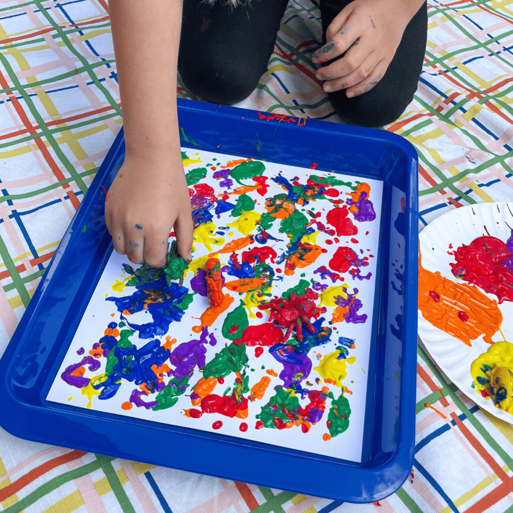 A child is painting with bugs by holding a green plastic grasshopper toy that had been dipped in green paint and presses it onto a white paper. The white paper sits in a shallow, blue tray. 