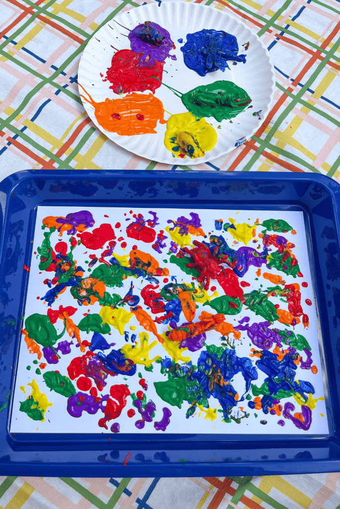 A paper plate with colorful puddles of paint and insect toys of matching colors sits next to a blue tray. In the blue tray is a white piece of paper covered in colorful paint and plastic bugs.