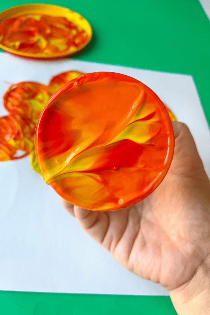 The bottom of a plastic cup that has been dipped in red, orange, and yellow paint is held up to show the colors swirled together.