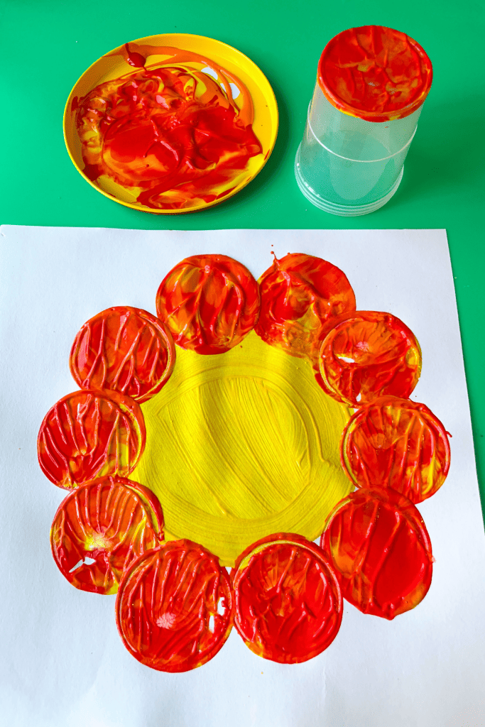 A white paper with "The Tiny Seed" flower art - a yellow circle painted in the center of a white paper and stamps made from the painty cup bottom dipped in paint around the yellow circle. 