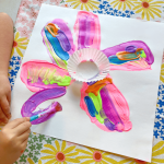 A child paints flower petals on her Cupcake Liner Flower Art (a white paper with a white cupcake liner glued to the middle and colorful petals painted around the cupcake liner center).