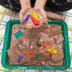 A preschool child cups her hands together, holding a handful of brown oobleck with plastic insect toys in it. Below her is a Frozen Oobleck Insect Sensory Bin.