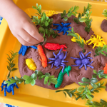 A child's hand pushes some faux greenery into Insect Garden Play Dough: a brown batch of play dough with faux greenery and plastic insect toys in it.
