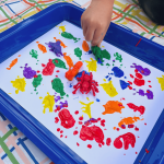A child is painting with bugs by holding a green plastic grasshopper toy that had been dipped in green paint and presses it onto a white paper. The white paper sits in a shallow, blue tray.