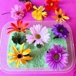 The spring garden insta-snow sensory bin (fluffy green faux "snow") sits on a pink table with faux flowers scattered around.