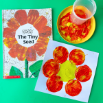 Eric Carle's book "The Tiny Seed" sits on a green able. Next to is a small yellow plate with yellow, red, and orange paint mixed together and a plastic cup sitting in the middle of the painty plate. Below is a white paper with "The Tiny Seed" flower art - a yellow circle painted in the center of a white paper and stamps made from the painty cup bottom dipped in paint around the yellow circle.