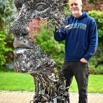 Brian Mock Standing Next To Human Face Sculpture Made Out Of Scrap Metal