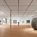 A large museum gallery with a large tire sculpture, a sculpture of silver figures dancing, and five paintings on the walls.