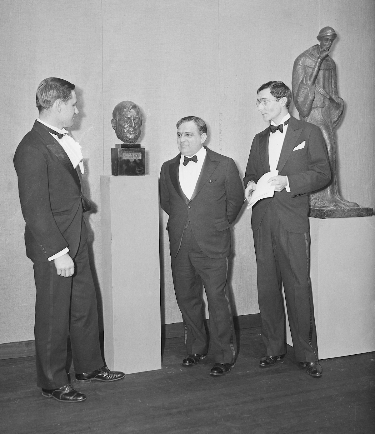 A black and white picture of three men in tuxedoes standing next to two sculptures.